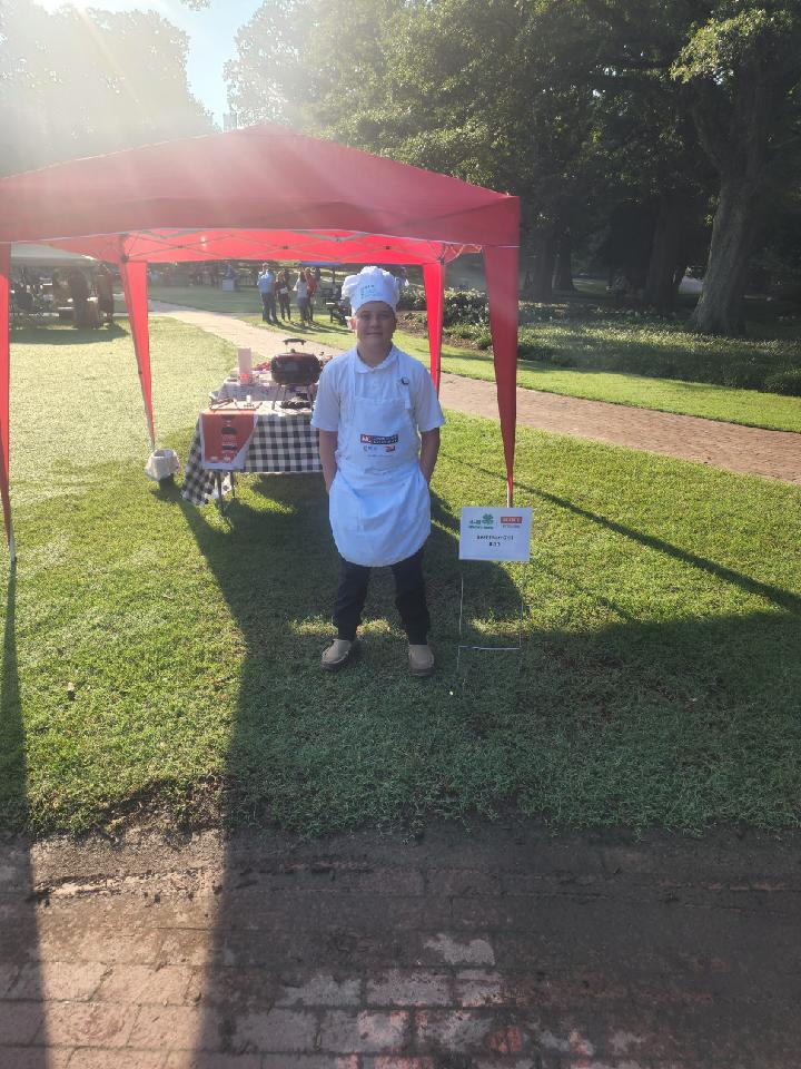 A boy dressed as a chef stands in front of a tent that contains cooking equipment.