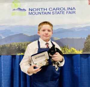 A youth poses in a dress shirt and blue apron while holding an award and his rabbit.