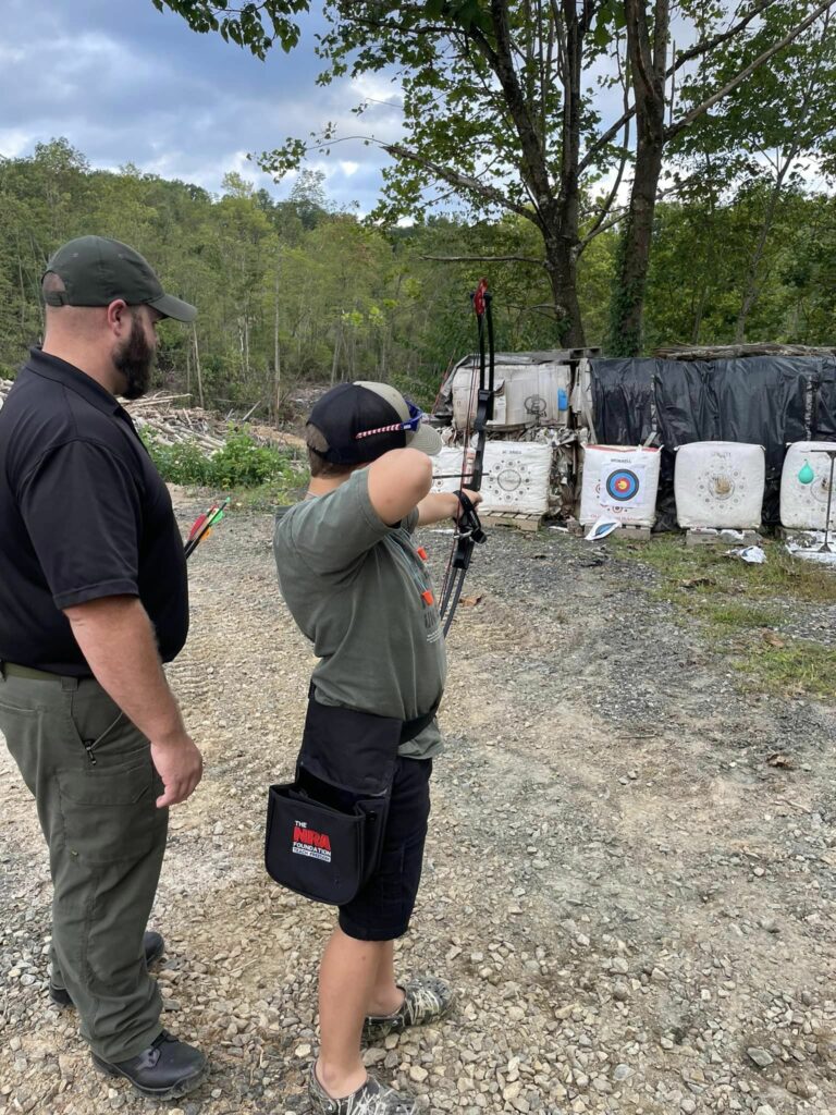 A youth aims a bow at a nearby target while an adult monitors.